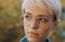 A person with short blonde hair and large glasses looks thoughtfully to the side. The background is a soft, blurred mix of natural colors.