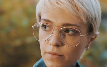 A person with short blonde hair and large glasses looks thoughtfully to the side. The background is a soft, blurred mix of natural colors.