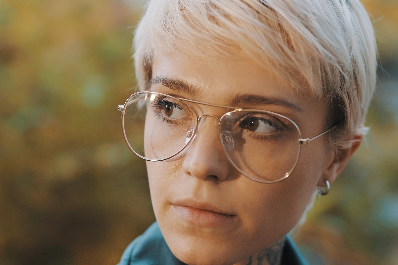 A person with short blonde hair and large glasses looks thoughtfully to the side. The background is a soft, blurred mix of natural colors.