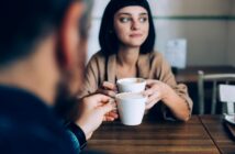 A woman with short dark hair holds a coffee cup, looking thoughtfully to the side while sitting at a wooden table. In the foreground, a blurred person holds another cup, suggesting a conversation between the two.