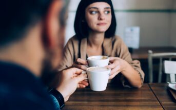 A woman with short dark hair holds a coffee cup, looking thoughtfully to the side while sitting at a wooden table. In the foreground, a blurred person holds another cup, suggesting a conversation between the two.