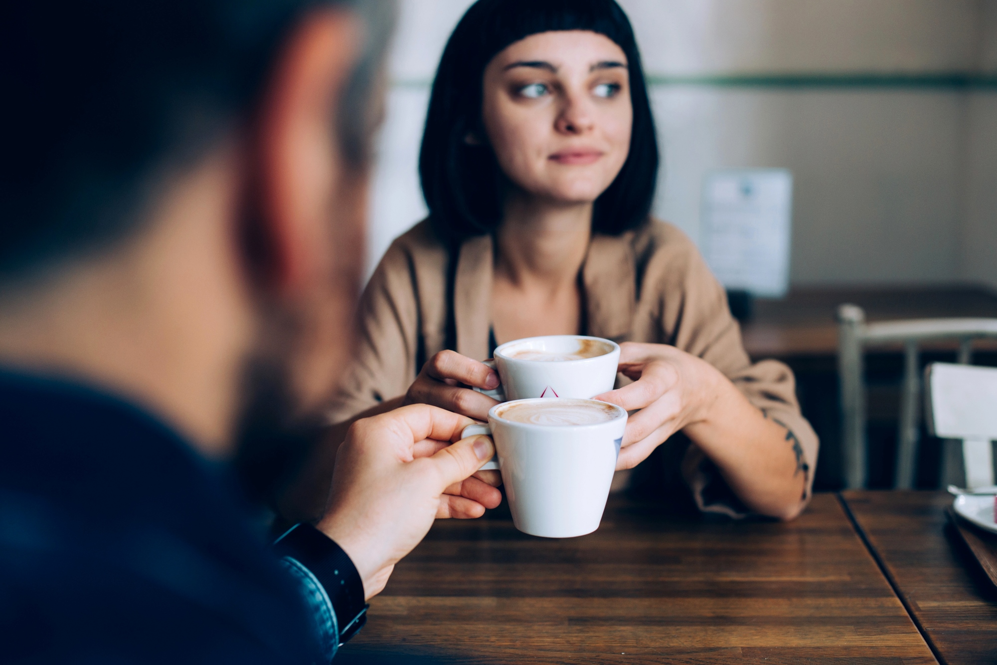 A woman with short dark hair holds a coffee cup, looking thoughtfully to the side while sitting at a wooden table. In the foreground, a blurred person holds another cup, suggesting a conversation between the two.