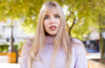 A woman with long blonde hair and bangs, wearing a light purple turtleneck sweater, stands outdoors. She has a surprised expression. The background features green trees and an out-of-focus urban setting.