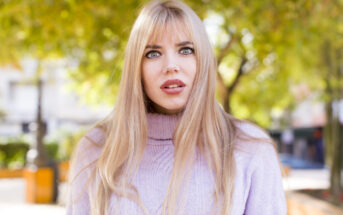 A woman with long blonde hair and bangs, wearing a light purple turtleneck sweater, stands outdoors. She has a surprised expression. The background features green trees and an out-of-focus urban setting.