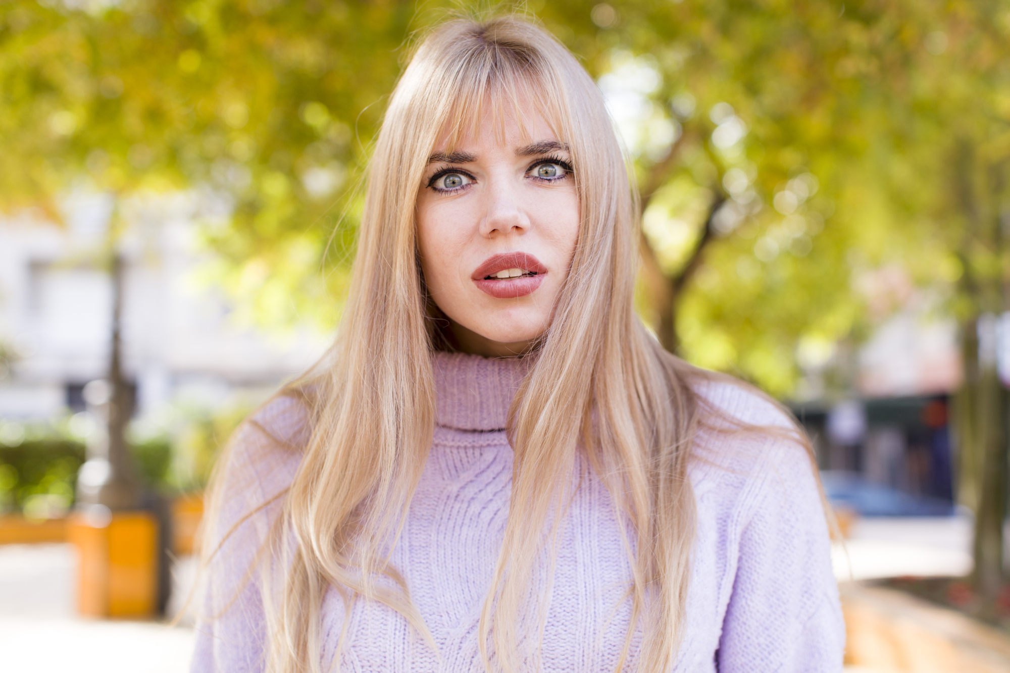 A woman with long blonde hair and bangs, wearing a light purple turtleneck sweater, stands outdoors. She has a surprised expression. The background features green trees and an out-of-focus urban setting.