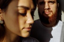 A close-up of two people with intense shadows across their faces. The person in the foreground has closed eyes, and the person in the background has a beard and directs their gaze at the camera. Both wear white shirts.