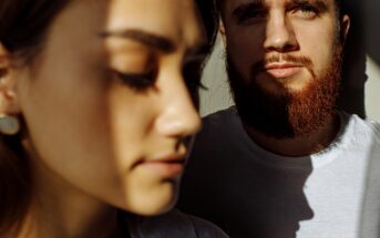 A close-up of two people with intense shadows across their faces. The person in the foreground has closed eyes, and the person in the background has a beard and directs their gaze at the camera. Both wear white shirts.