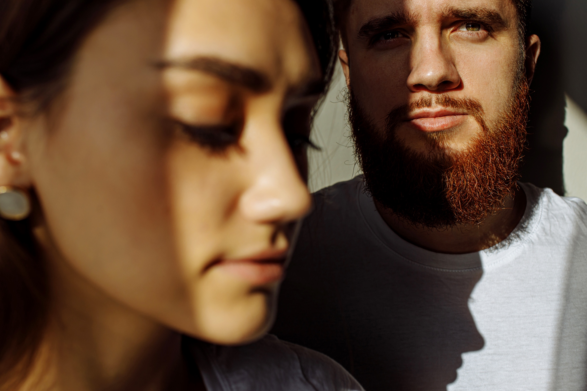 A close-up of two people with intense shadows across their faces. The person in the foreground has closed eyes, and the person in the background has a beard and directs their gaze at the camera. Both wear white shirts.