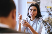 A woman with short dark hair, wearing hoop earrings and a light sweater, is sitting and holding a mug. She is gesturing with her other hand, pointing upwards, and appears to be engaged in conversation with a person blurred in the foreground.