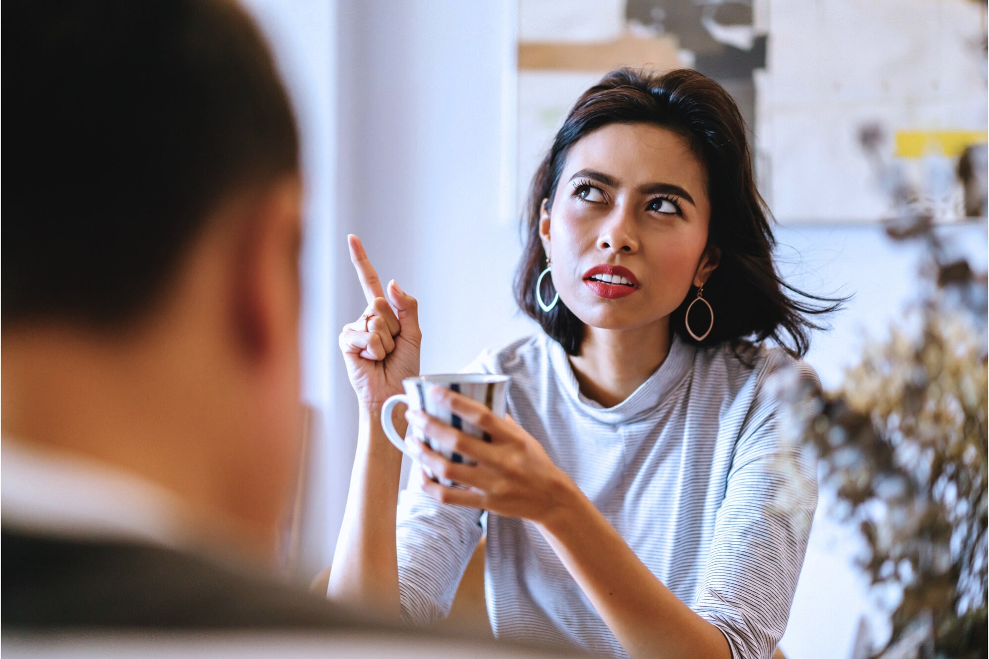 A woman with short dark hair, wearing hoop earrings and a light sweater, is sitting and holding a mug. She is gesturing with her other hand, pointing upwards, and appears to be engaged in conversation with a person blurred in the foreground.