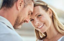 A couple stands close together, the man's forehead touching the woman's. The woman is smiling warmly, her long blond hair blowing slightly. The background is blurred, suggesting an outdoor setting in soft, natural light.