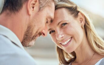 A couple stands close together, the man's forehead touching the woman's. The woman is smiling warmly, her long blond hair blowing slightly. The background is blurred, suggesting an outdoor setting in soft, natural light.