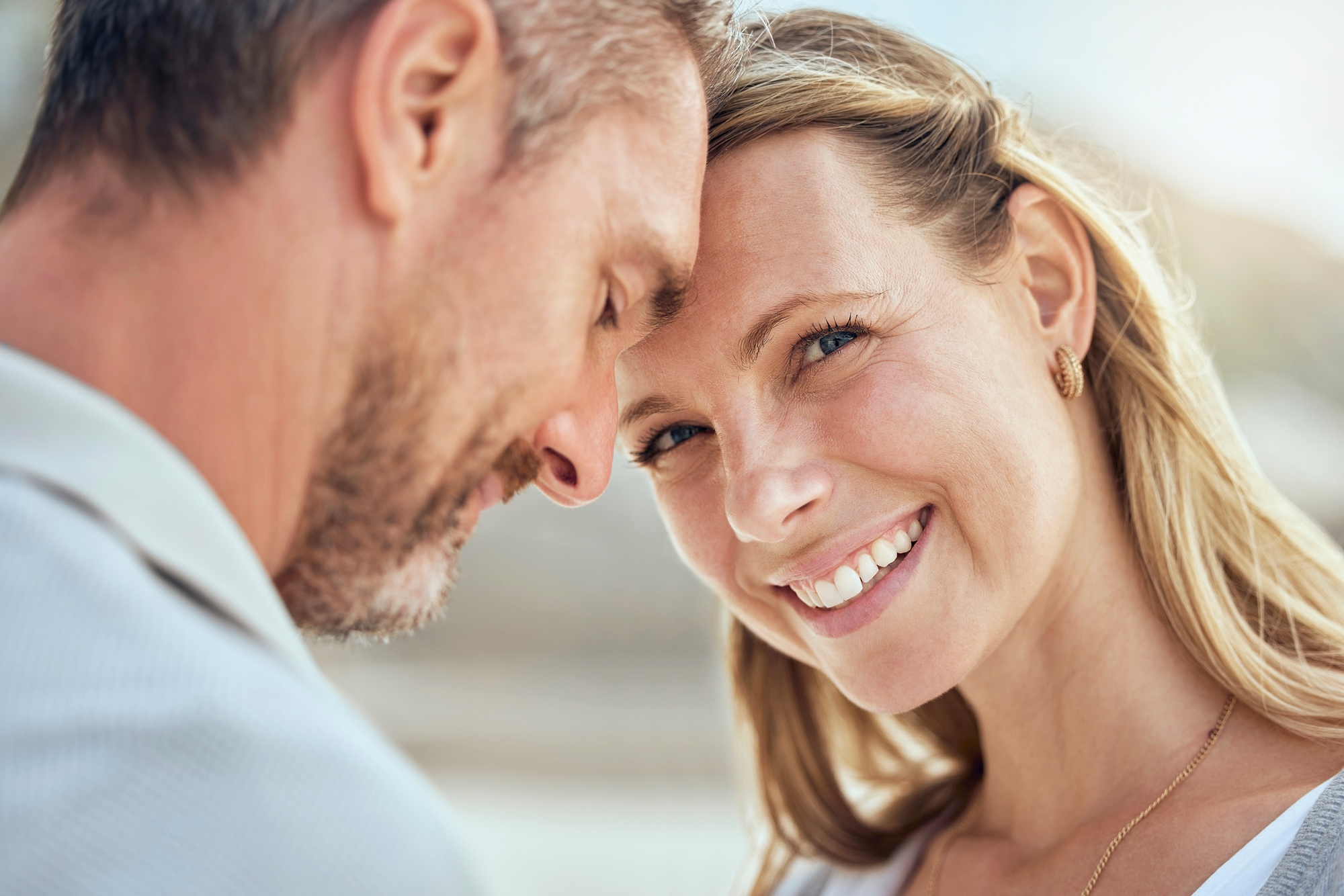 A couple stands close together, the man's forehead touching the woman's. The woman is smiling warmly, her long blond hair blowing slightly. The background is blurred, suggesting an outdoor setting in soft, natural light.