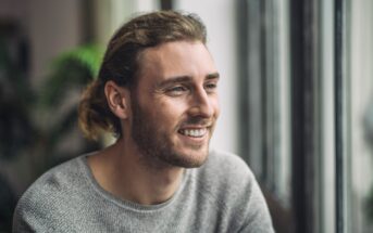 A person with long, light brown hair smiles while looking out of a window. They're wearing a gray sweater, with a background of blurred greenery and bright natural light.