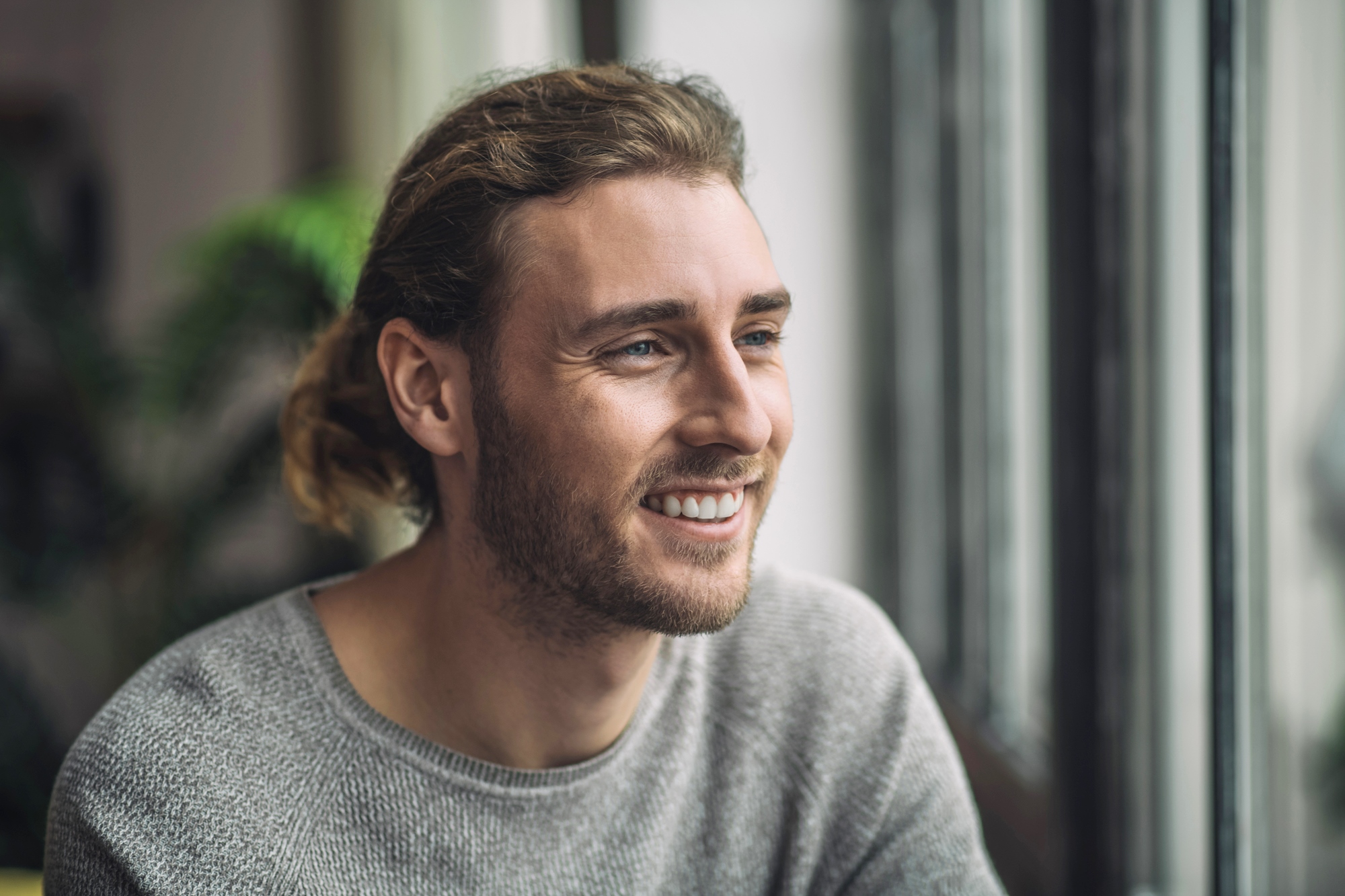 A person with long, light brown hair smiles while looking out of a window. They're wearing a gray sweater, with a background of blurred greenery and bright natural light.