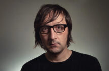 A person with shoulder-length brown hair and black-rimmed glasses, wearing a black shirt, is looking directly at the camera against a neutral background.