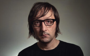 A person with shoulder-length brown hair and black-rimmed glasses, wearing a black shirt, is looking directly at the camera against a neutral background.