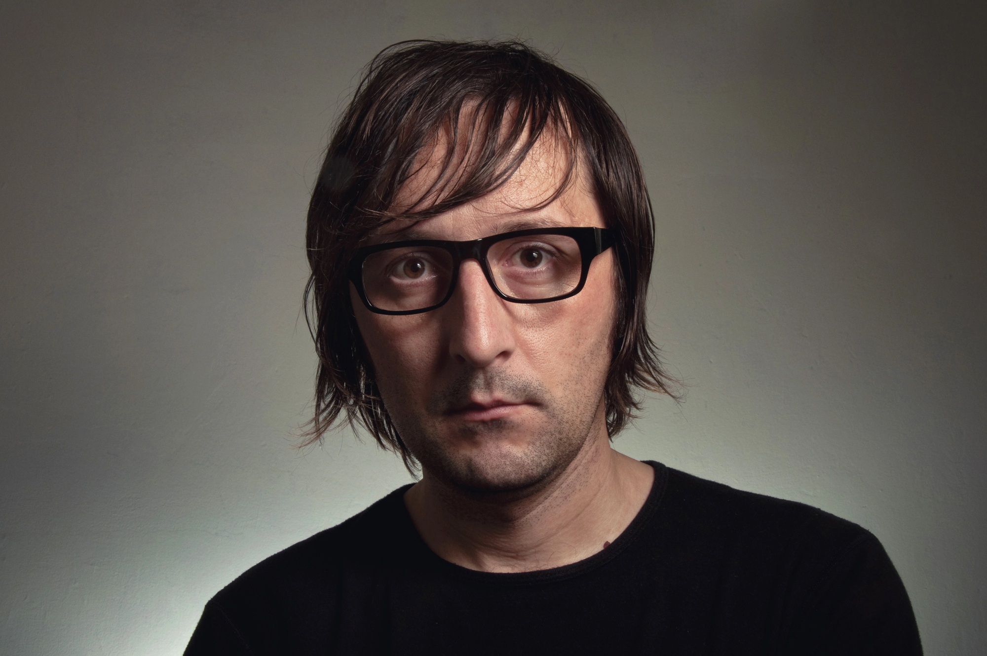 A person with shoulder-length brown hair and black-rimmed glasses, wearing a black shirt, is looking directly at the camera against a neutral background.