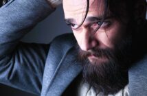 A bearded man with long hair looks down thoughtfully, wearing a grey jacket over a white sweater. A strand of hair falls across his face as he rests his hand on his head in a dimly lit environment.