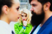 A woman in a bright green coat, looking concerned and biting her finger, stands between a man with a beard and another woman in the foreground, who are out of focus. The image suggests a tense or dramatic situation.