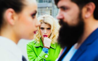 A woman in a bright green coat, looking concerned and biting her finger, stands between a man with a beard and another woman in the foreground, who are out of focus. The image suggests a tense or dramatic situation.