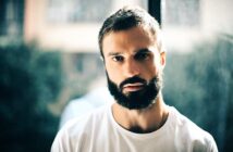 Man with a thick beard and short hair wearing a white t-shirt, gazing intently at the camera. He is standing indoors with a softly blurred background and natural lighting coming from a window behind him.