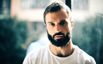 Man with a thick beard and short hair wearing a white t-shirt, gazing intently at the camera. He is standing indoors with a softly blurred background and natural lighting coming from a window behind him.