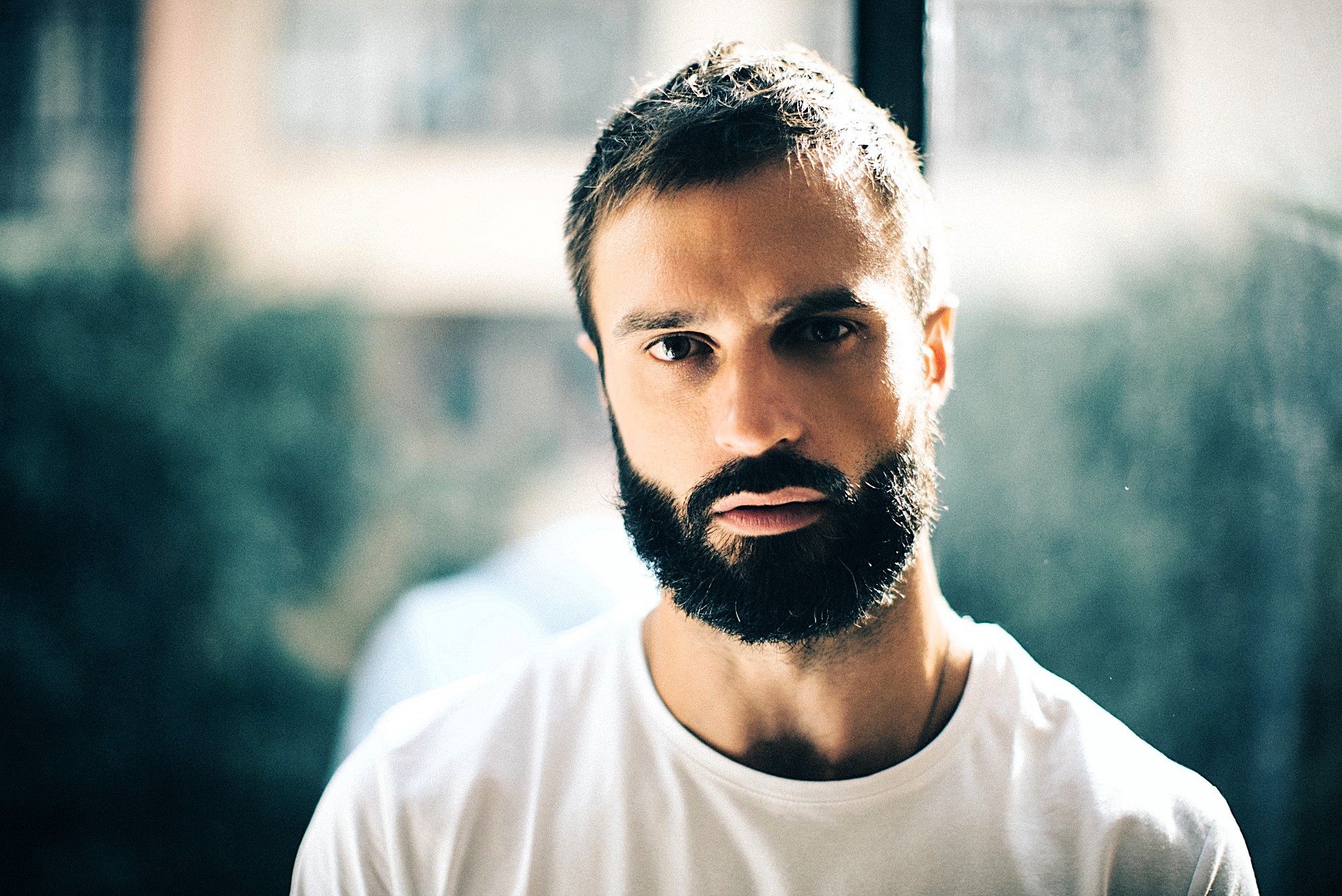 Man with a thick beard and short hair wearing a white t-shirt, gazing intently at the camera. He is standing indoors with a softly blurred background and natural lighting coming from a window behind him.