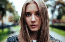 A young woman with long brown hair stands outdoors. She is wearing a dark jacket, and her expression is neutral. The background features a blurred park-like setting with trees and a pathway.