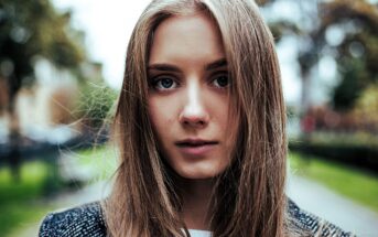 A young woman with long brown hair stands outdoors. She is wearing a dark jacket, and her expression is neutral. The background features a blurred park-like setting with trees and a pathway.