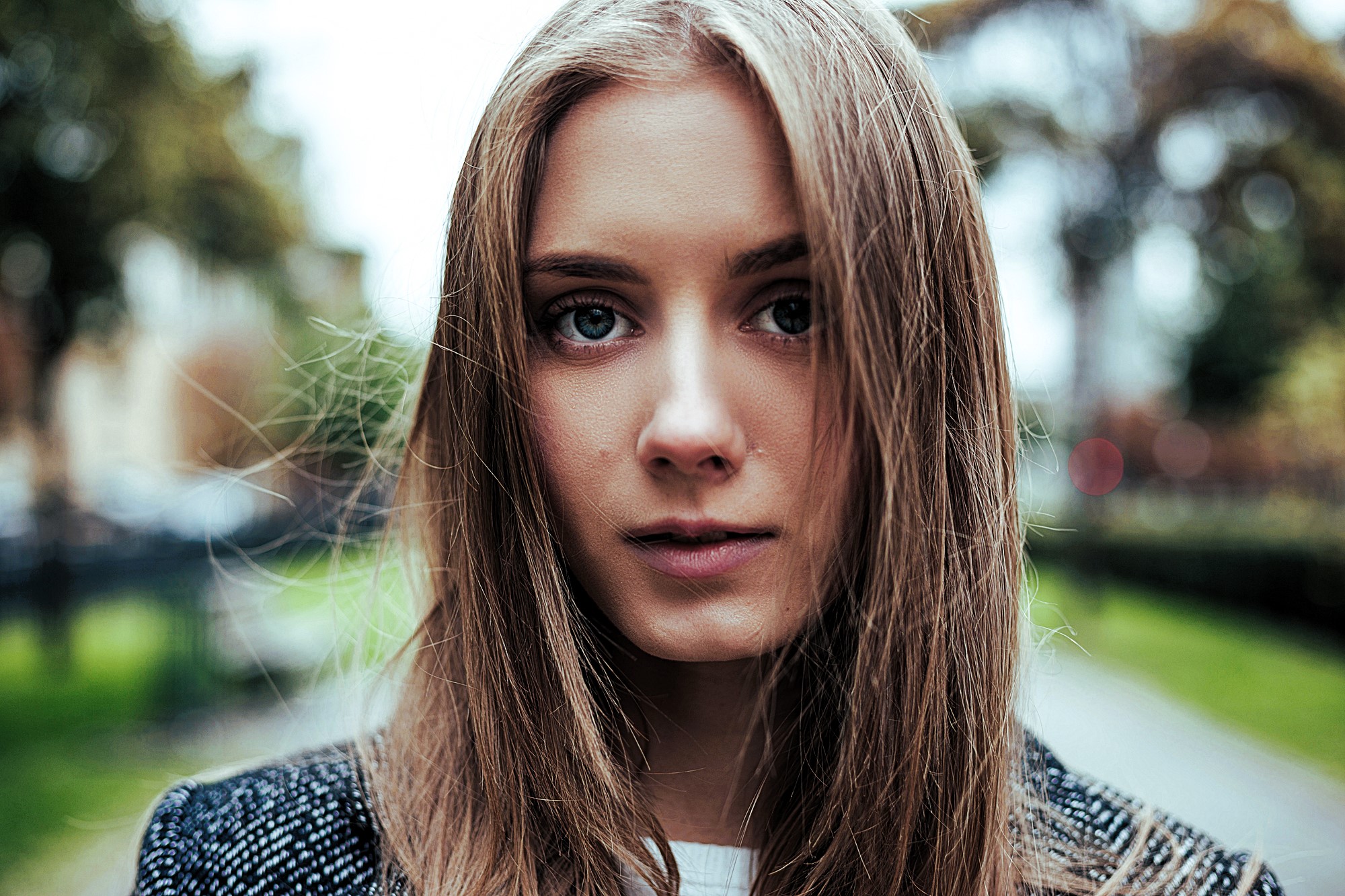 A young woman with long brown hair stands outdoors. She is wearing a dark jacket, and her expression is neutral. The background features a blurred park-like setting with trees and a pathway.