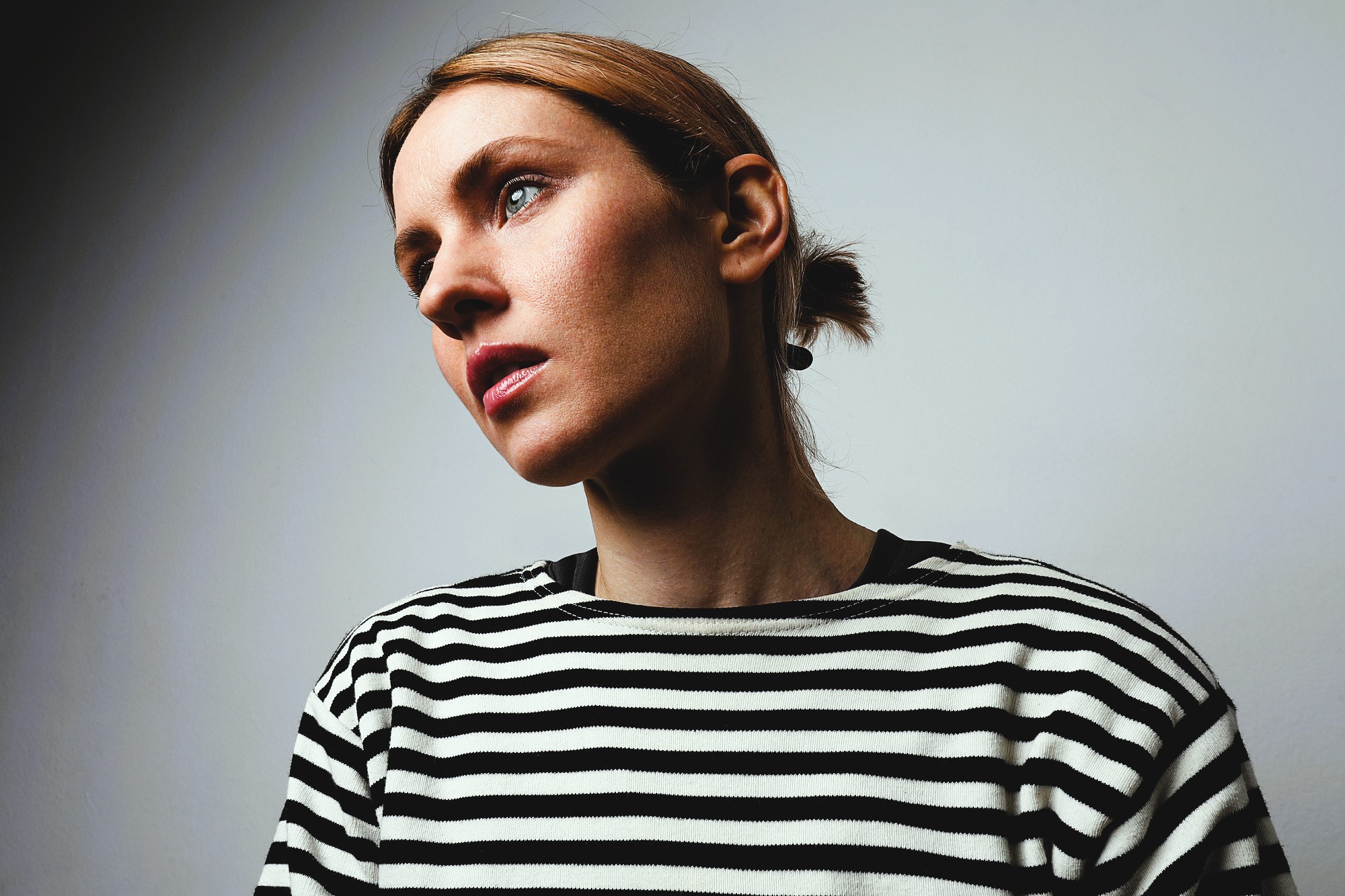 A person with light brown hair is looking to the side against a plain background. They are wearing a black and white striped shirt.