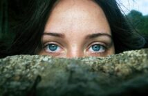 A close-up of a person with striking blue eyes, partially hidden behind a rock. Dark hair frames their face, creating a mysterious atmosphere as they gaze directly at the viewer.