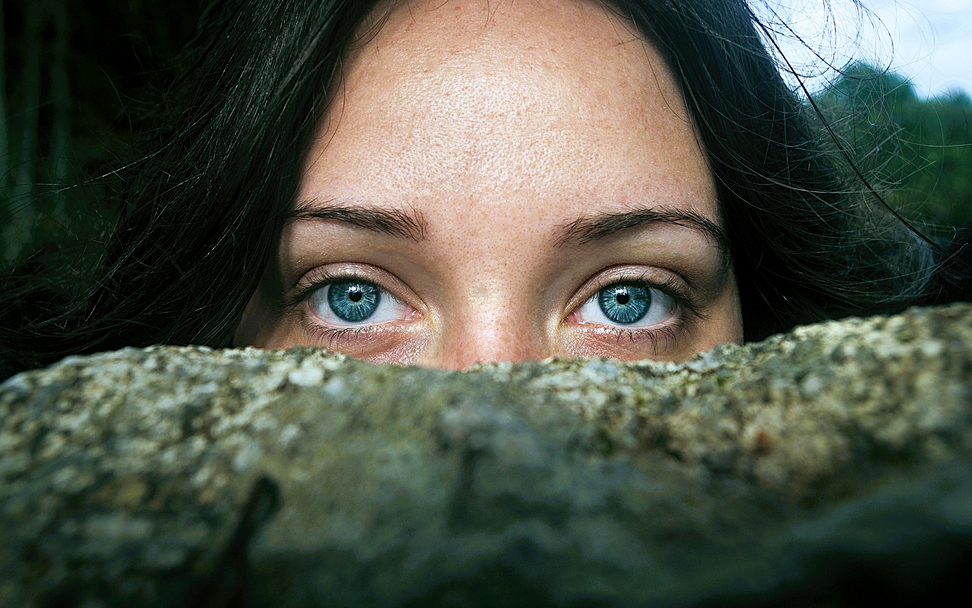 A close-up of a person with striking blue eyes, partially hidden behind a rock. Dark hair frames their face, creating a mysterious atmosphere as they gaze directly at the viewer.