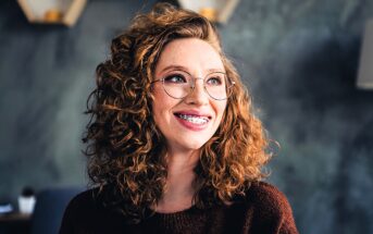 A person with curly red hair and glasses smiles, looking off to the side. They are wearing a dark sweater, and the background is blurred, featuring a muted, textured wall with hexagonal shelves.