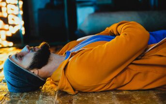 A bearded man wearing an orange hoodie and a gray beanie lies on a wooden floor, facing upward, with his hands clasped over his chest. The background is dimly lit with warm, ambient lights.