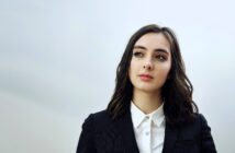 A person with shoulder-length dark hair wearing a black blazer over a white shirt stands against a plain background, looking thoughtfully to the side.