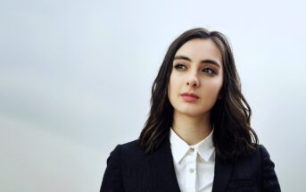 A person with shoulder-length dark hair wearing a black blazer over a white shirt stands against a plain background, looking thoughtfully to the side.