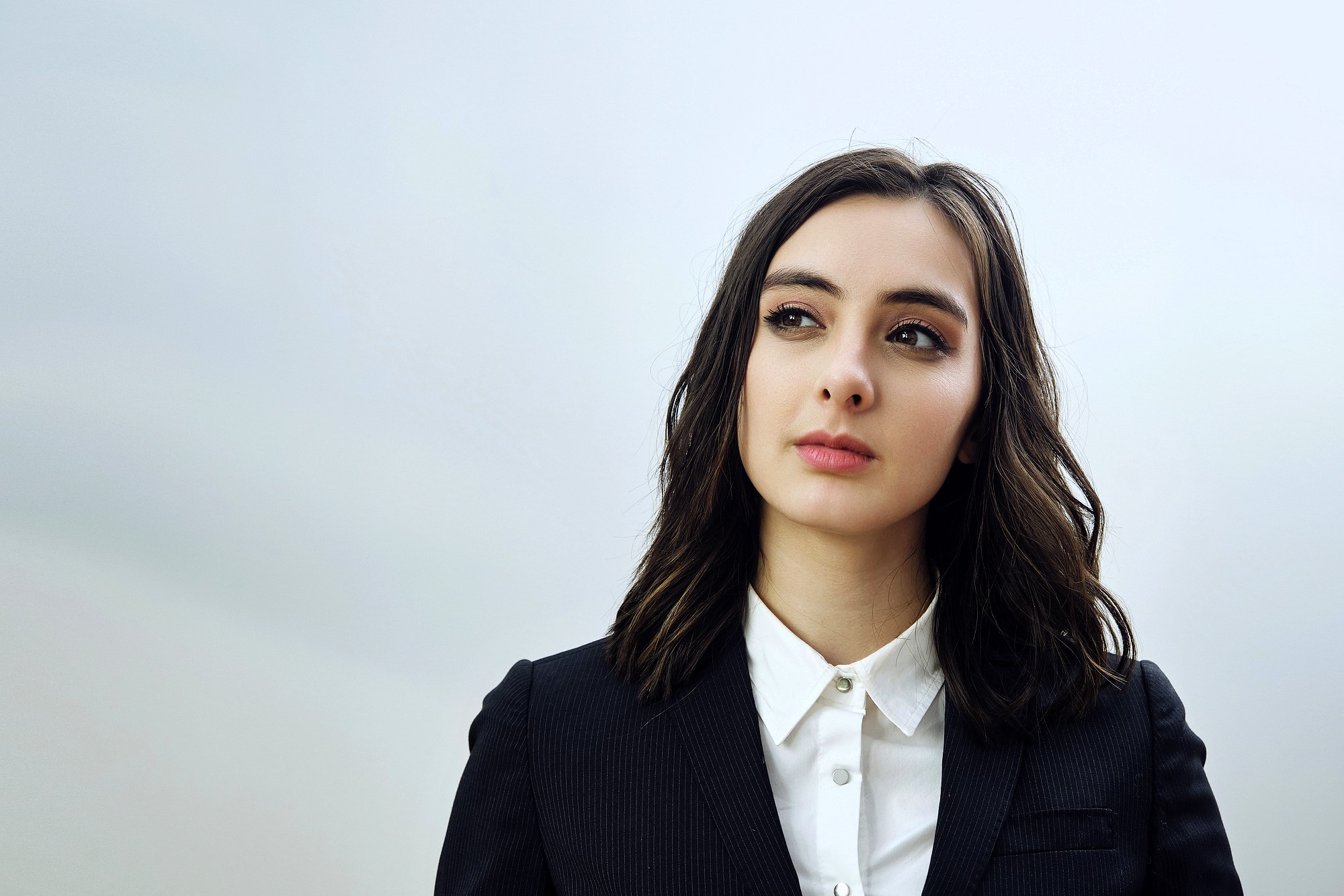 A person with shoulder-length dark hair wearing a black blazer over a white shirt stands against a plain background, looking thoughtfully to the side.