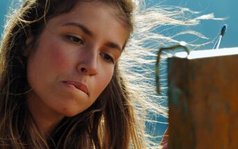 A person with long, windblown hair and a nose ring is concentrating on painting outdoors. They are focused, with brushes in hand, working on a canvas against a blurred background.