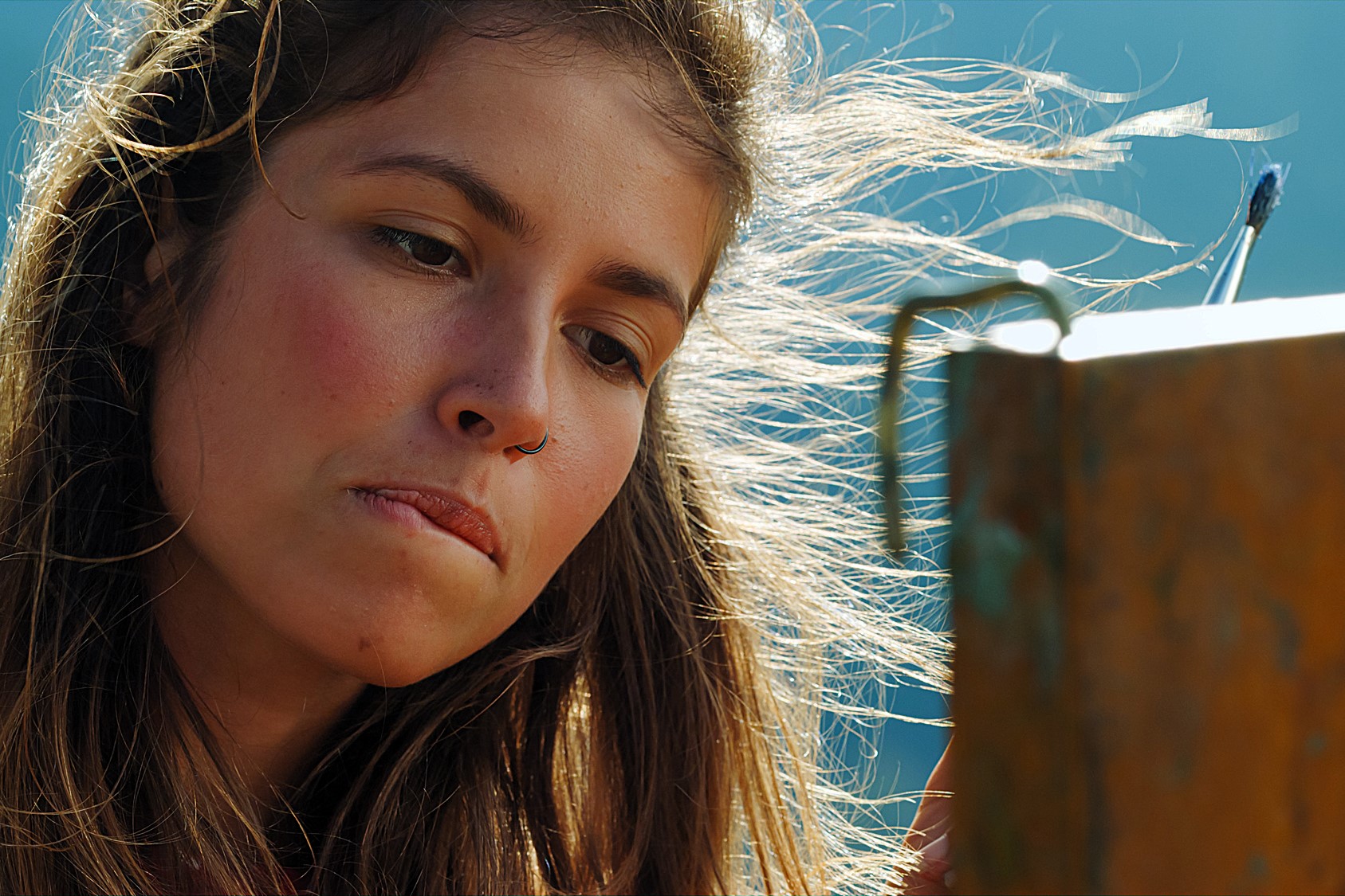 A person with long, windblown hair and a nose ring is concentrating on painting outdoors. They are focused, with brushes in hand, working on a canvas against a blurred background.