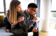 A woman with long hair comforts a man sitting on a gray couch. The man, wearing glasses and a sweater, appears thoughtful or upset. They are in a modern, well-lit room with large windows in the background.