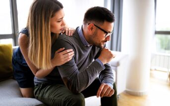 A woman with long hair comforts a man sitting on a gray couch. The man, wearing glasses and a sweater, appears thoughtful or upset. They are in a modern, well-lit room with large windows in the background.