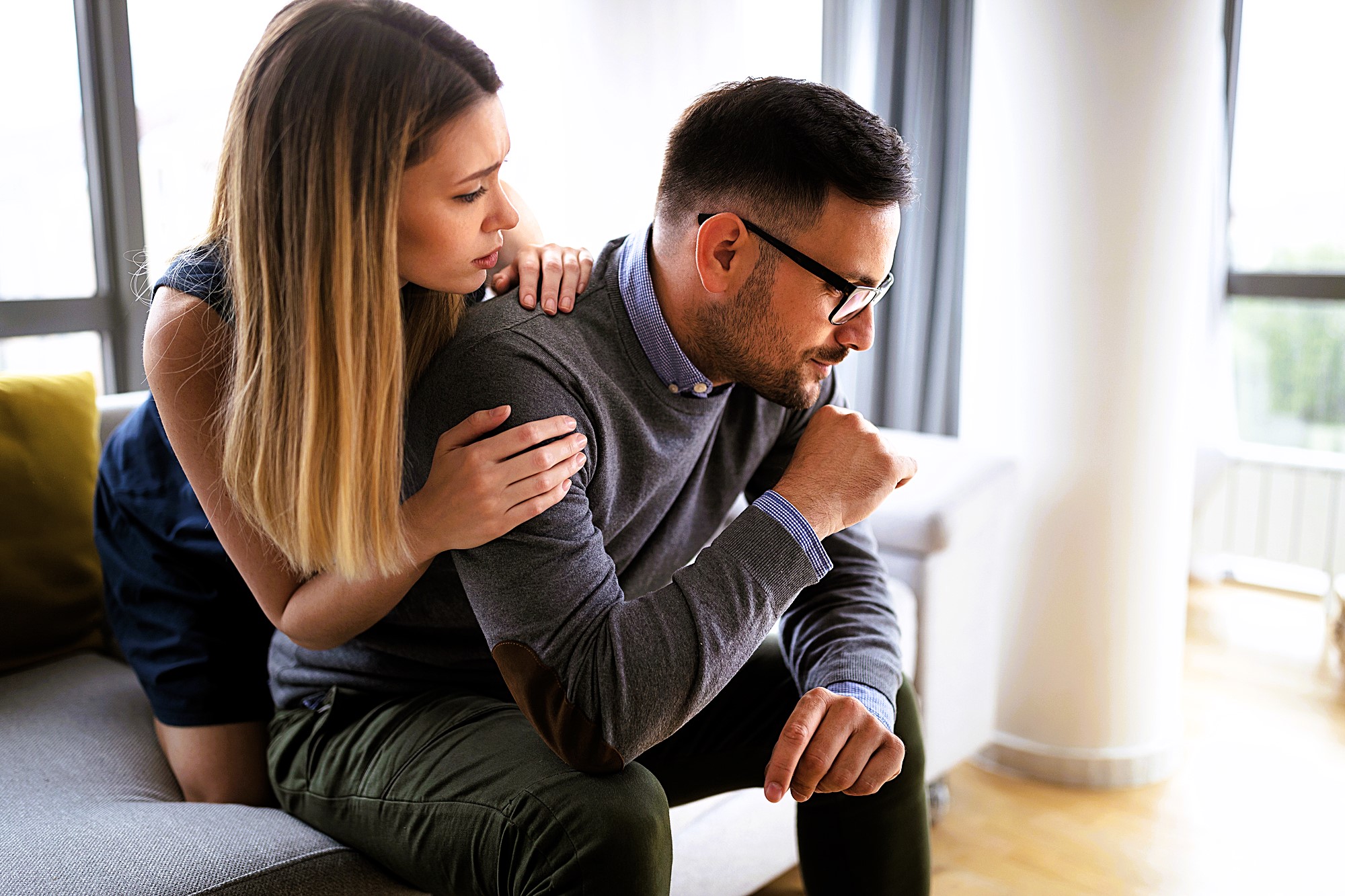 A woman with long hair comforts a man sitting on a gray couch. The man, wearing glasses and a sweater, appears thoughtful or upset. They are in a modern, well-lit room with large windows in the background.