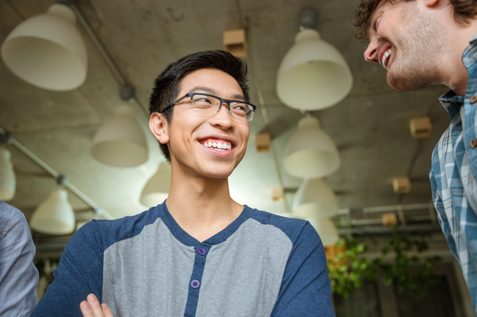 A young man wearing glasses and a blue shirt is smiling and looking up in a brightly lit room with hanging light fixtures. Another person is partially visible, also smiling, in the foreground.