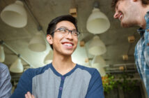 A smiling person with glasses, wearing a blue and gray shirt, is looking up. Another person is partially visible, also smiling. They are indoors, with large, hanging lights and some greenery in the background.