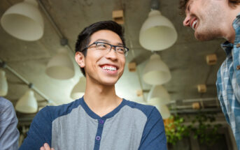 A smiling person with glasses, wearing a blue and gray shirt, is looking up. Another person is partially visible, also smiling. They are indoors, with large, hanging lights and some greenery in the background.