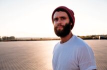 A man with a beard wears a red beanie and white t-shirt, standing in an open area with a paved surface. The sun is setting in the background, casting a warm glow over the scene.