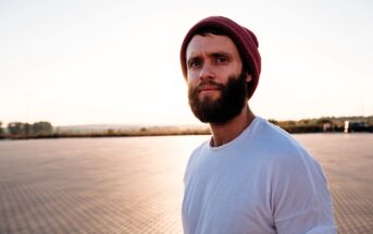 A man with a beard wears a red beanie and white t-shirt, standing in an open area with a paved surface. The sun is setting in the background, casting a warm glow over the scene.