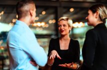 Three people are engaged in a conversation indoors. A woman with short blonde hair is speaking and gesturing with her hands while a man and woman listen attentively. The background features blurred lights and glass walls.